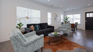 A spacious living room with a dark grey sofa, patterned armchairs, a round coffee table, and a dining area with a patterned tablecloth.