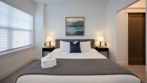 Hotellihuone, jossa on suuri sänky, kaksi yöpöytää valaisimineen, taulu seinällä ja ovi oikealla puolella.