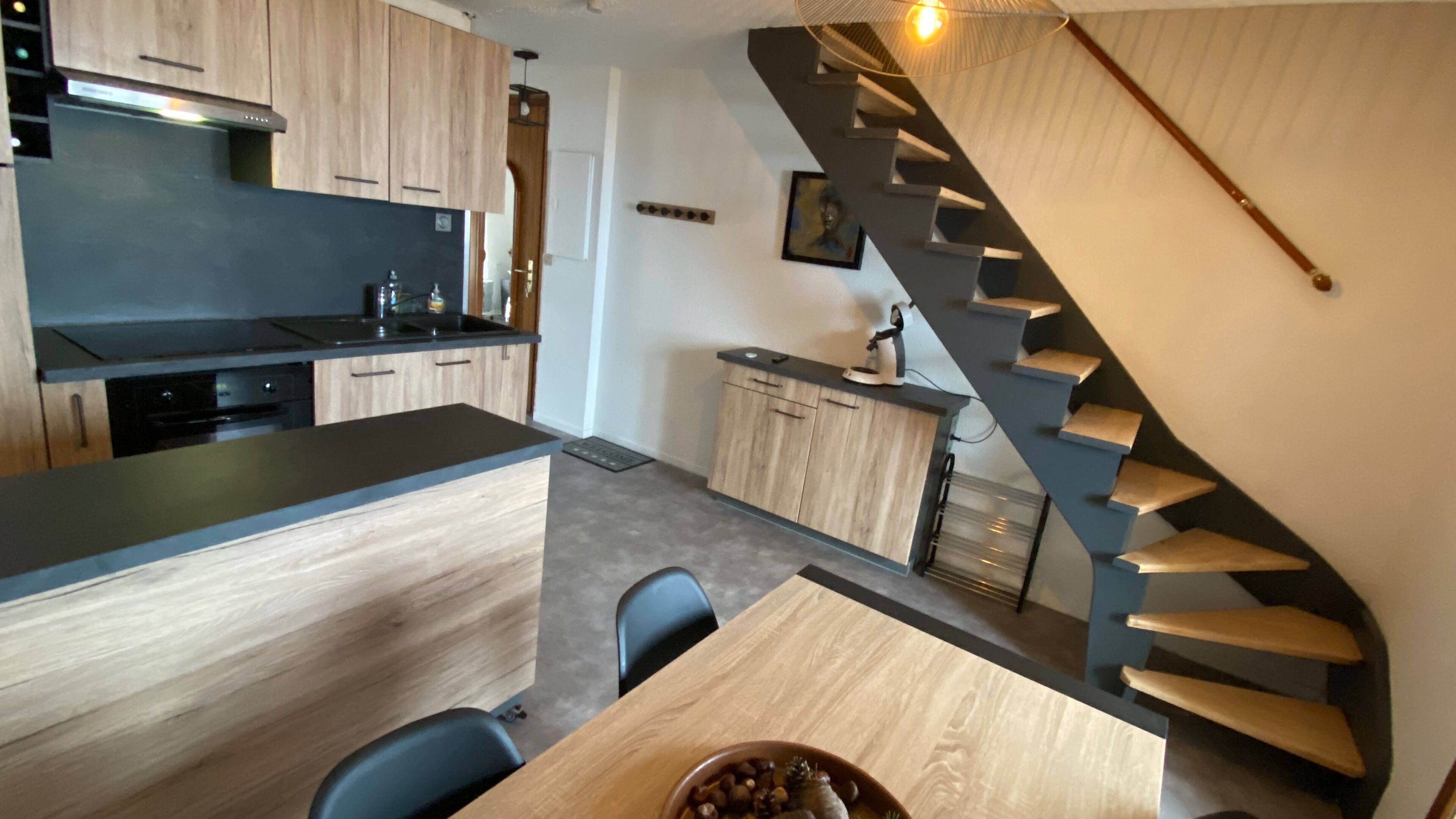 Een moderne keuken met houten kastjes, een zwart aanrecht en een ingebouwde oven. Er is een eethoek met een tafel en stoelen, en een spiltrap die naar een andere verdieping leidt.