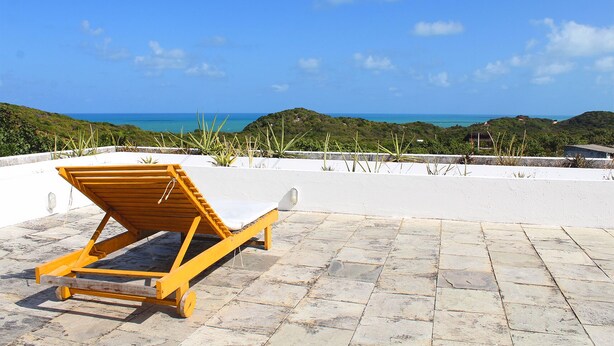 A wooden lounge chair on a stone patio with a view of the ocean and hills. - Toca do Sol com vista natureza em Pipa (Tibau do Sul)