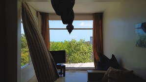 A room with a large window, a sofa, a chair, and a hanging light fixture. - Toca do Sol com vista natureza em Pipa (Tibau do Sul)