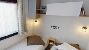 A small, well-lit room with two beds, a nightstand, and a shelf with books and a decorative item. - Mobile home from 2022 at La Carabasse campsite (Vias)