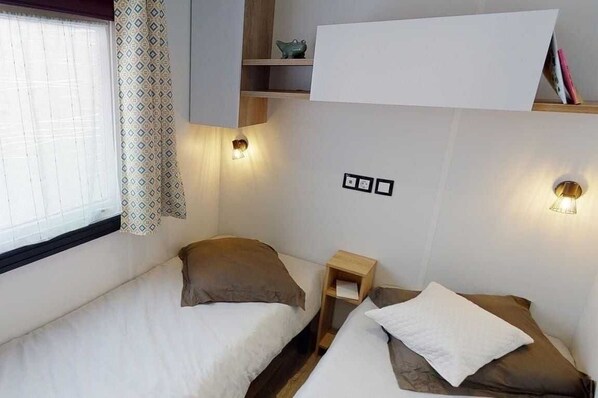 Una habitación pequeña y luminosa con dos camas, una mesita de noche y un estante con libros y un objeto decorativo.