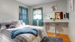A bedroom with a bed, a desk with two stools, a mirror, and artwork on the wall.