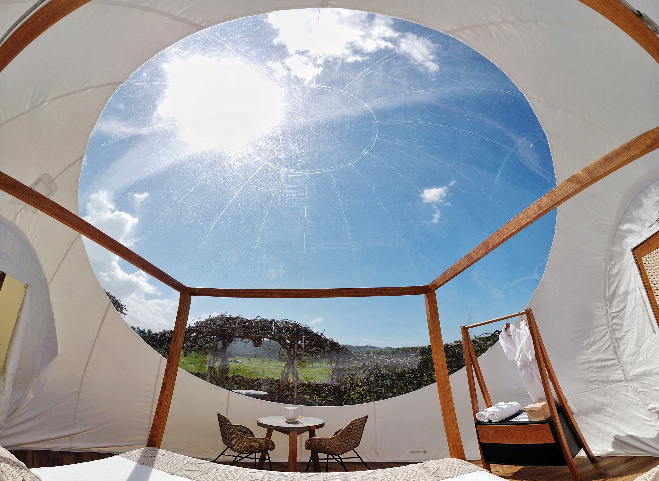 A dome-shaped tent with a clear view of a grassy landscape and a tree. Inside, there is a small table with chairs and a bed.