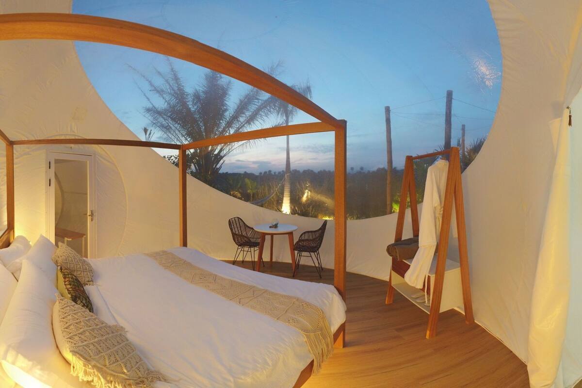 Een tent met een bed, een tafel en stoelen, en uitzicht op bomen en de lucht.