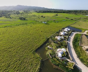 Vista aérea de un paisaje rural con campos verdes, un río y un conjunto de estructuras en forma de cúpula a lo largo de un camino de tierra.