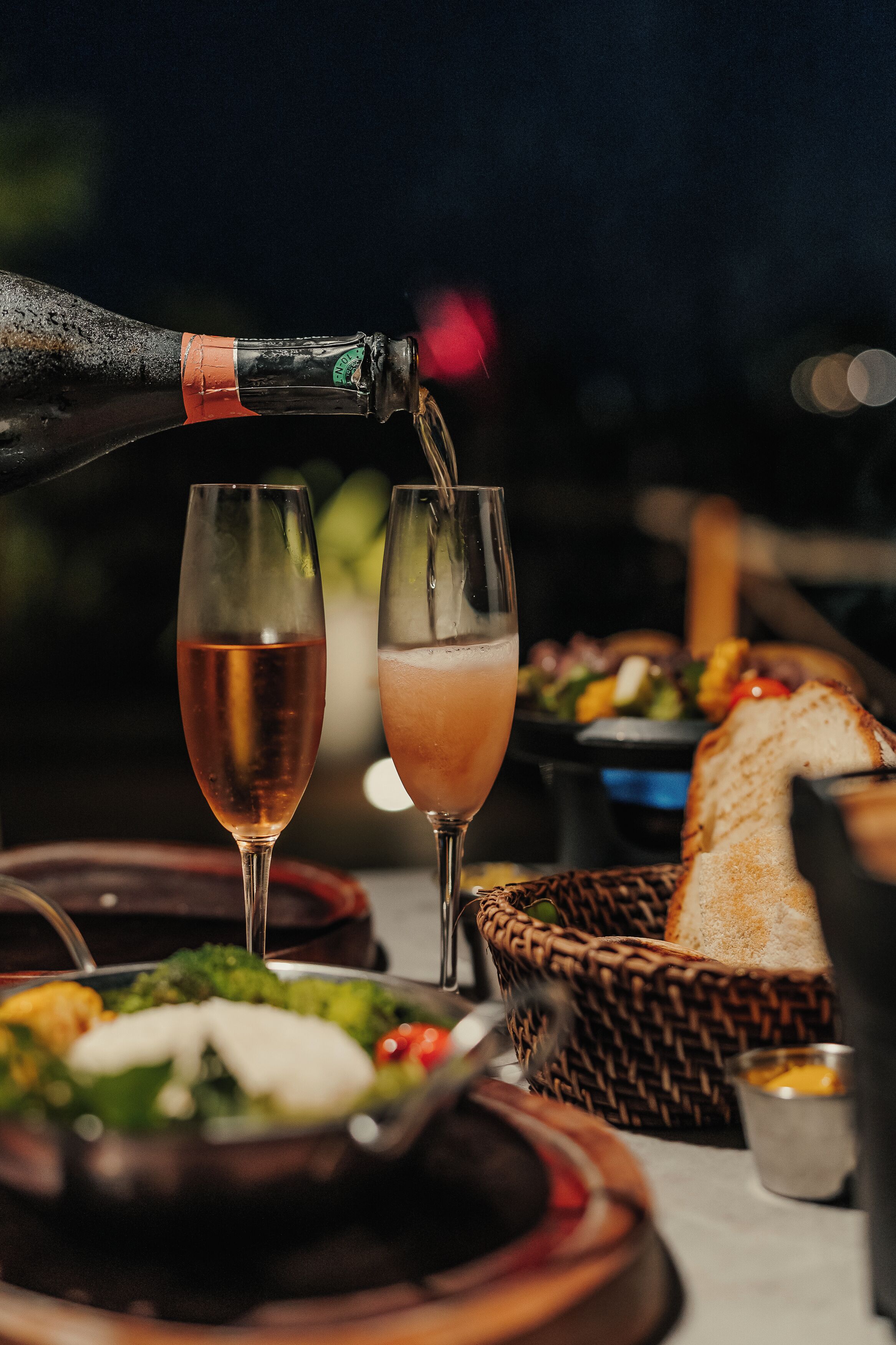 A bottle of champagne being poured into two glasses, with a basket of bread and a plate of food in the background.