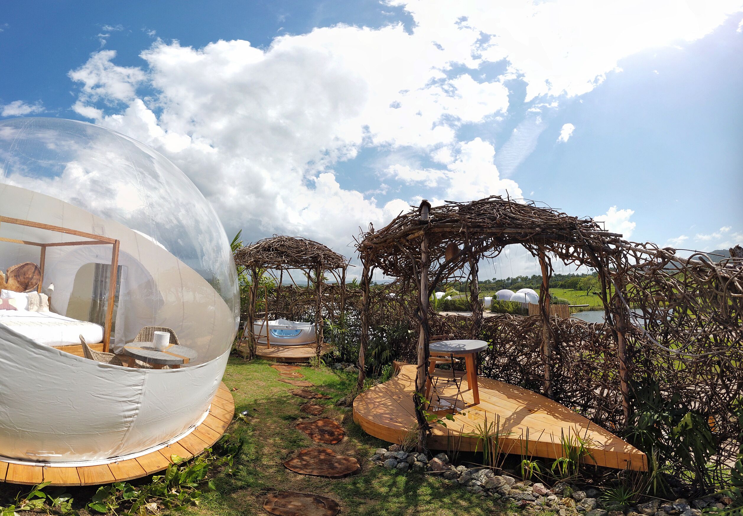 A transparent dome structure with a bed and seating area, surrounded by a garden with a wooden deck and a gazebo.