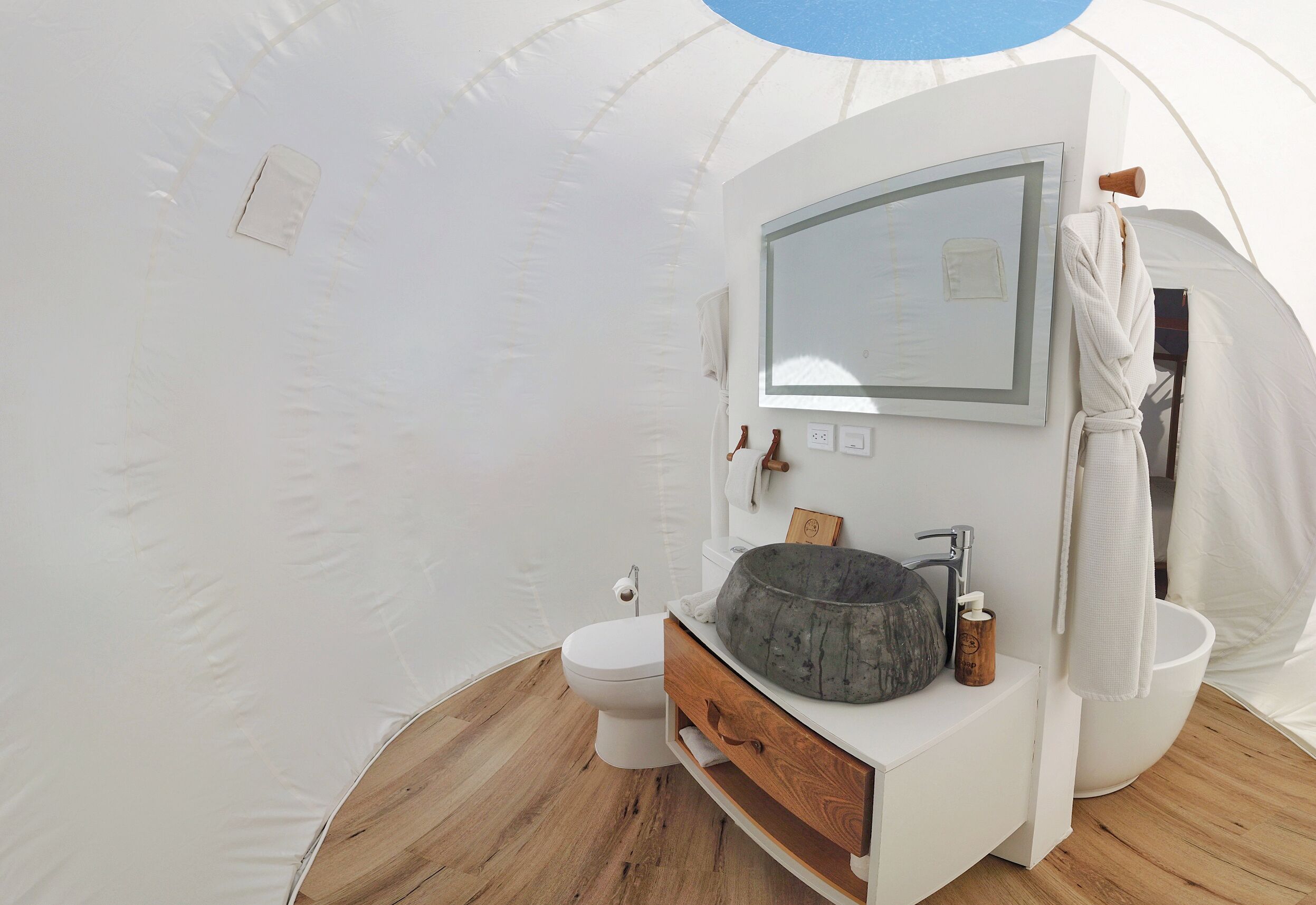 Una carpa con interior blanco, suelo de madera, inodoro blanco, tocador de madera con lavamanos y un cuadro en la pared.