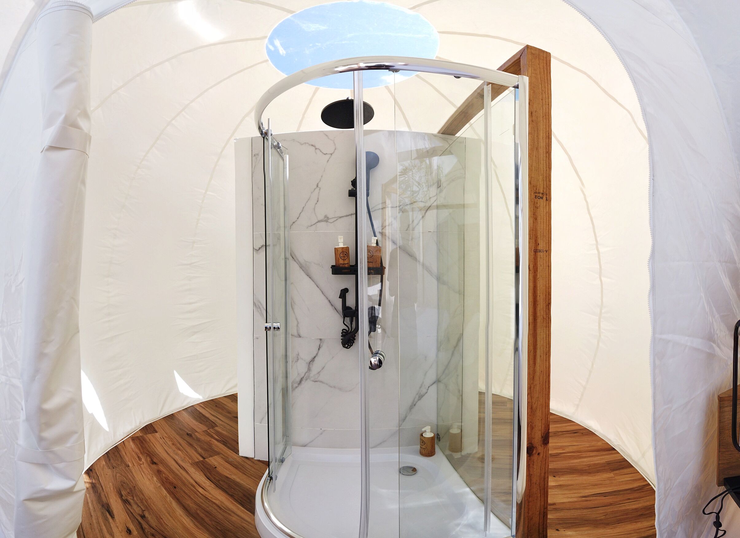 A glass-enclosed shower with a wooden floor, a marble wall, and a wooden shelf with toiletries.