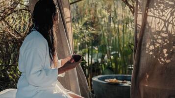 A person sitting on a bed in a rustic outdoor setting with a table and a teapot.