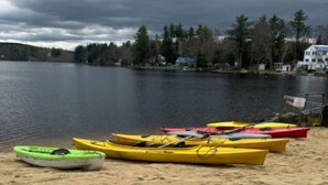 A sandy beach with several kayaks lined up, a calm lake in the background, and a cloudy sky. - Lakefront Pink Cottage (Northwood)