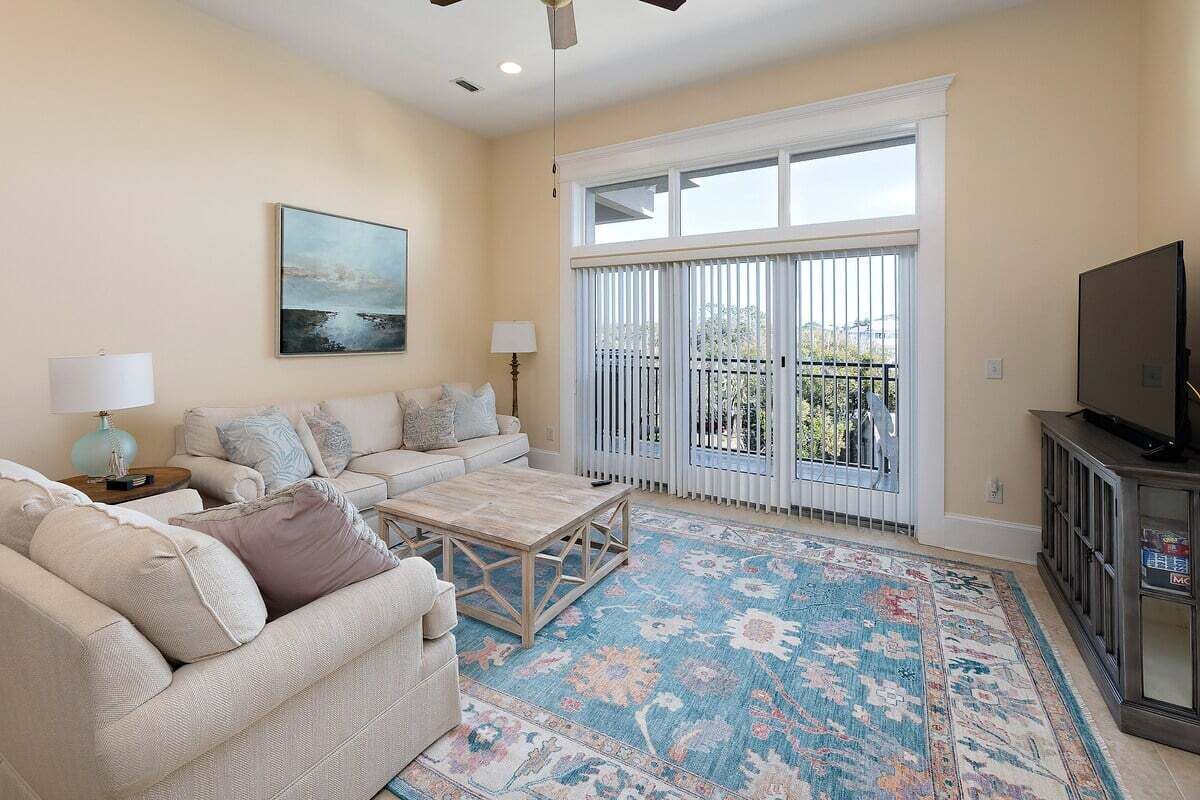 A living room with a beige sofa, a coffee table, a TV on a cabinet, a large window with blinds, and a painting on the wall.