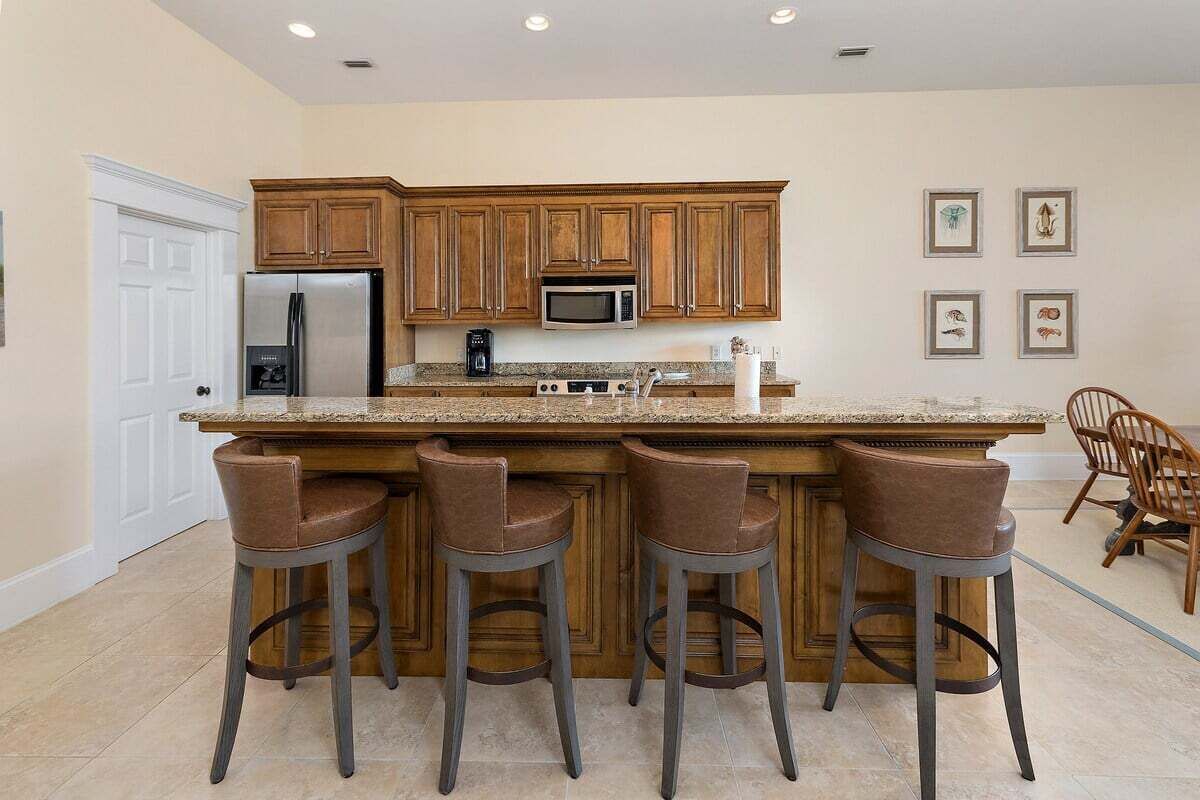 A kitchen with wooden cabinets, a stainless steel refrigerator, a microwave, and a granite countertop. There are four bar stools and a dining area with a wooden chair.