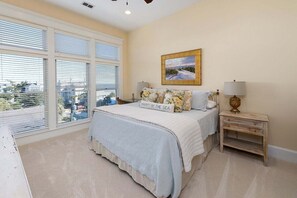 Un dormitorio con un ventanal grande, una cama con almohada decorada con motivos marinos, una mesita de noche con lámpara y un cuadro en la pared.