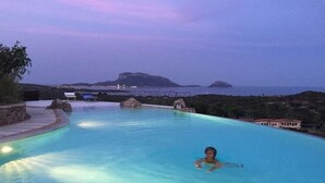 A swimming pool with a person in the water, surrounded by a stone deck and a view of the sea and hills. - Villa Smeraldo (Sottomonte)