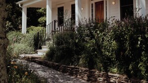 A white colonial-style house with a covered porch, stone pathway, and lush greenery.