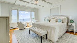 A bedroom with a large bed, a bench, a chair, a nightstand, and a view of the sea through large windows. - Ocean front and 3 beaches across,parks, short walk to historic downtown! (Gloucester)