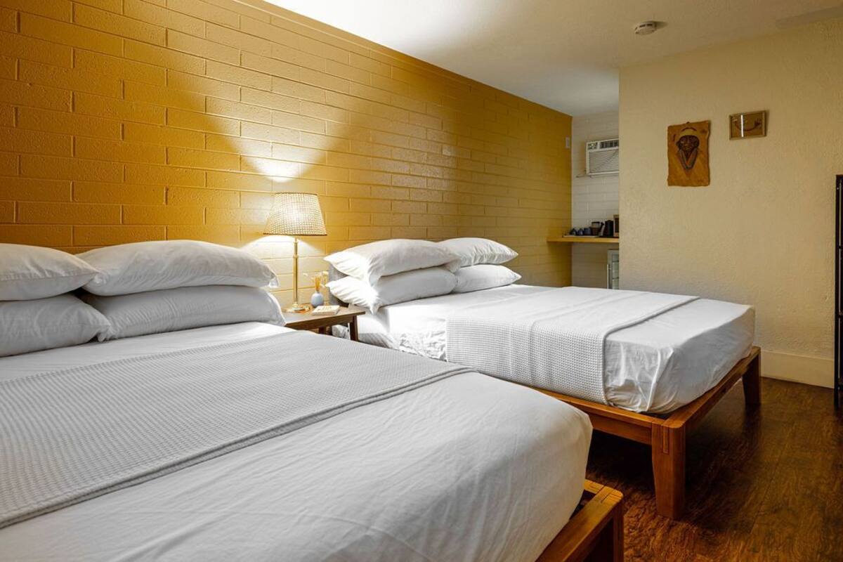 A room with two beds, a brick wall, a wooden floor, and a small table with a lamp.