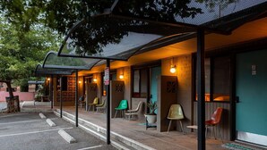 A covered outdoor seating area with chairs and a potted plant, a brick building with a green door, and a parking lot.