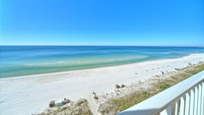 Blick auf einen Strand vom Balkon aus, mit blauem Himmel, Sandstrand und einigen Gebäuden am Ufer.