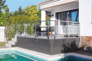 A modern outdoor pool area with a glass balustrade, a seating area with chairs, and a staircase leading to a covered patio. - Modern Villa close to Costabella beach (Marbella)