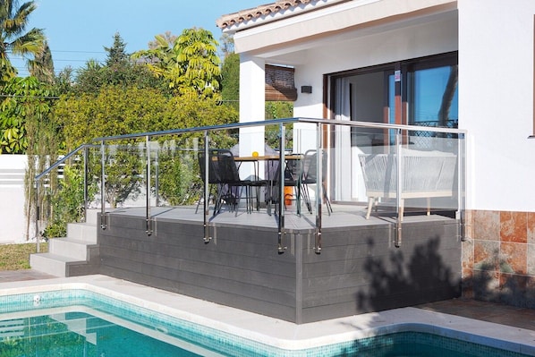 Un área de piscina moderna al aire libre con barandilla de vidrio, zona de estar con sillas y una escalera que conduce a un porche cubierto.