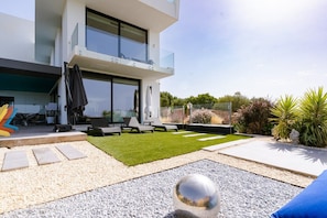 A modern outdoor patio with a glass balustrade, a grassy area, and a pebble path leading to a seating area with lounge chairs.