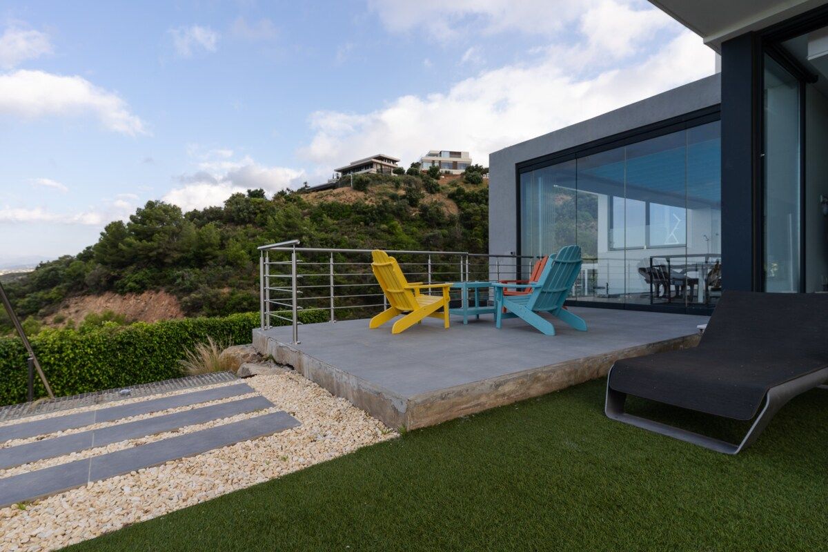 A modern outdoor terrace with a glass balustrade, a lounge chair, and two Adirondack chairs.