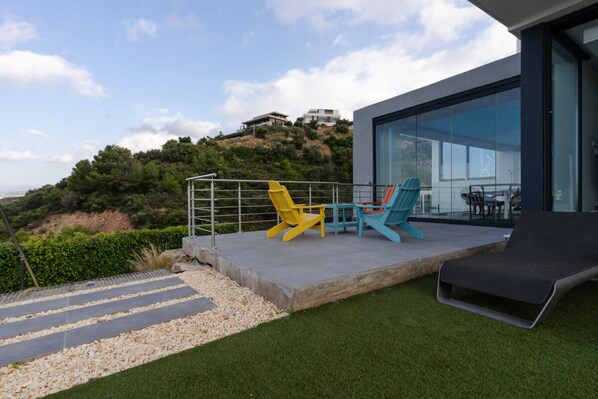 Une terrasse extérieure moderne dotée d’une balustrade en verre, d’un fauteuil de relaxation et de deux chaises Adirondack.