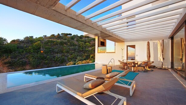 Un área de piscina moderna al aire libre con sillones de descanso y una mesa de comedor, con vista a una ladera.