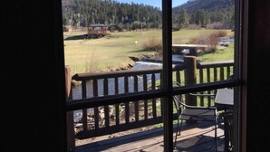Vista desde una ventana con terraza de madera, con vista a un río, un campo de césped y una colina boscosa.