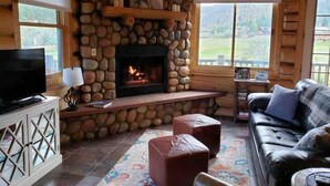 Interior acogedor de una cabaña de troncos con chimenea de piedra, un televisor sobre un mueble bajo, un sofá de cuero y vistas al exterior a través de amplios ventanales.