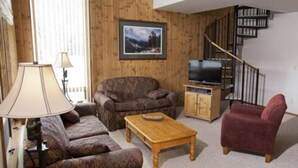 A living room with a sofa, armchair, and a wooden coffee table. There is a TV stand with a television and a cabinet. A staircase is visible in the background. - (1)Big Sky 2BR Condo w/outdoor heated pool/hot tub (Big Sky)