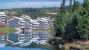 A residential building complex with multiple balconies, a lake in front, and a forested hillside in the background. - (1)Big Sky 2BR Condo w/outdoor heated pool/hot tub (Big Sky)