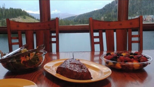 A table with a plate of food, a bowl of fruit, and a scenic view through large windows. - (1)Big Sky 2BR Condo w/outdoor heated pool/hot tub (Big Sky)
