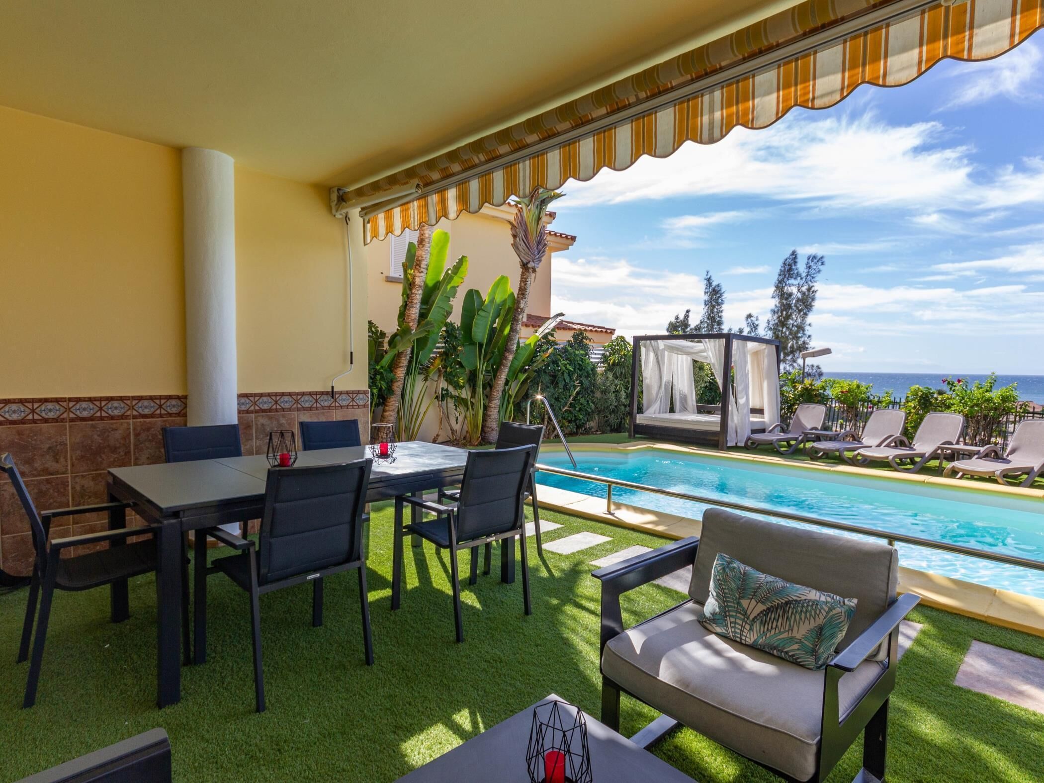 A poolside area with a table and chairs, a covered seating area, and a view of the ocean.
