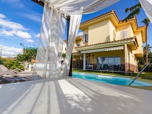 A poolside area with a clear view of a yellow building with a tiled roof, a covered outdoor seating area, and a swimming pool.