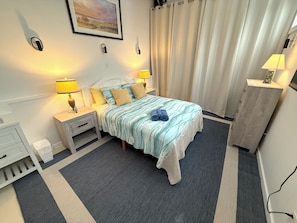 A neatly arranged hotel room with a bed, bedside lamps, a nightstand, a dresser, and a painting on the wall. - Beautiful Coastal Loft - 5 Minute Walk to Beach (Jamestown)
