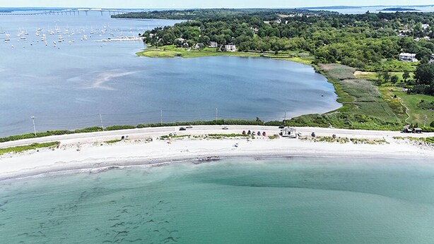 Oversigtsbillede af et kystområde med en sandstrand, en bro i baggrunden og en gruppe huse omgivet af grønt.