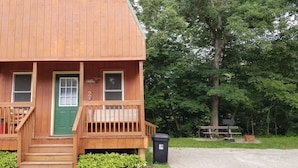 Eine kleine Holzhütte mit einer grünen Tür, zwei Fenstern, einer Veranda und einem Mülleimer.