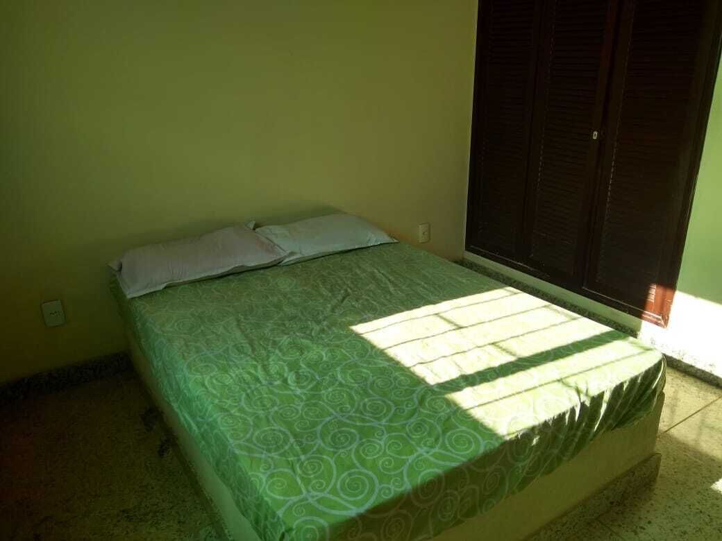 A single bed with a patterned bedspread in a room with a plain yellow wall and a window with closed blinds.