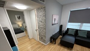 Un salon moderne avec un canapé noir, une petite table et un banc. Une porte donne accès à une chambre à coucher équipée d’un lit et d’une table de chevet. La pièce est dotée d’un plancher en bois et d’un mur d’accent en briques grises.