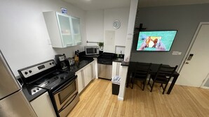 Une cuisine compacte équipée d’un réfrigérateur, d’une cuisinière, d’un four à micro-ondes et de placards. On y trouve une table à manger avec des chaises, et un téléviseur fixé au mur.