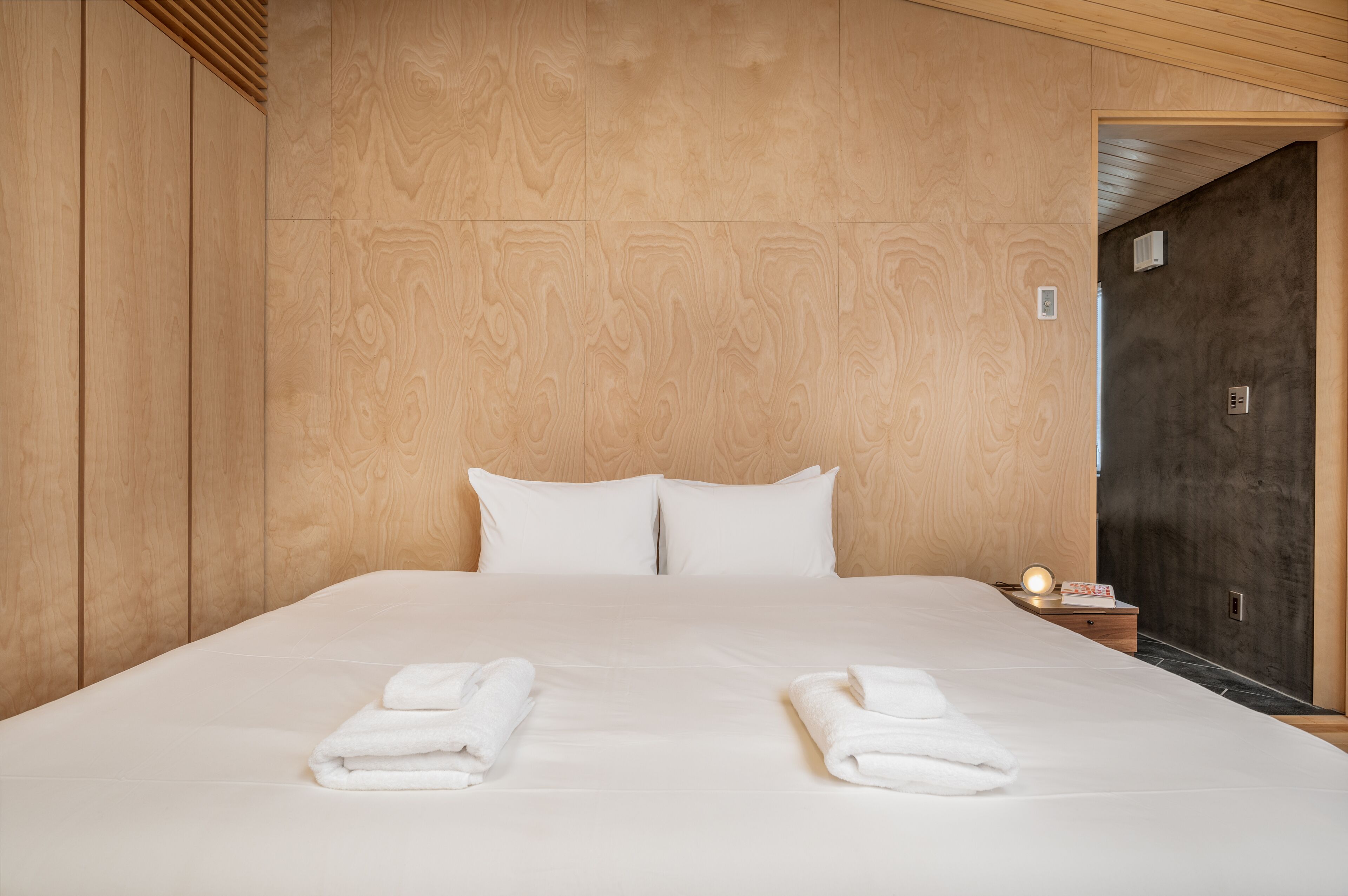 A neatly made bed with white linens and two folded towels on the side, a bedside table with a lamp, and a wooden headboard.