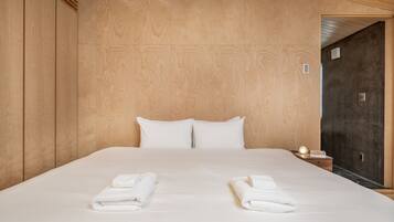 A neatly made bed with white linens and two folded towels on the side, a bedside table with a lamp, and a wooden headboard.