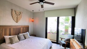 Een slaapkamer met een bed, een plafondventilator, een macramé decoratie aan de muur, een nachtkastje, een stoel, een televisie en een balkon met uitzicht.