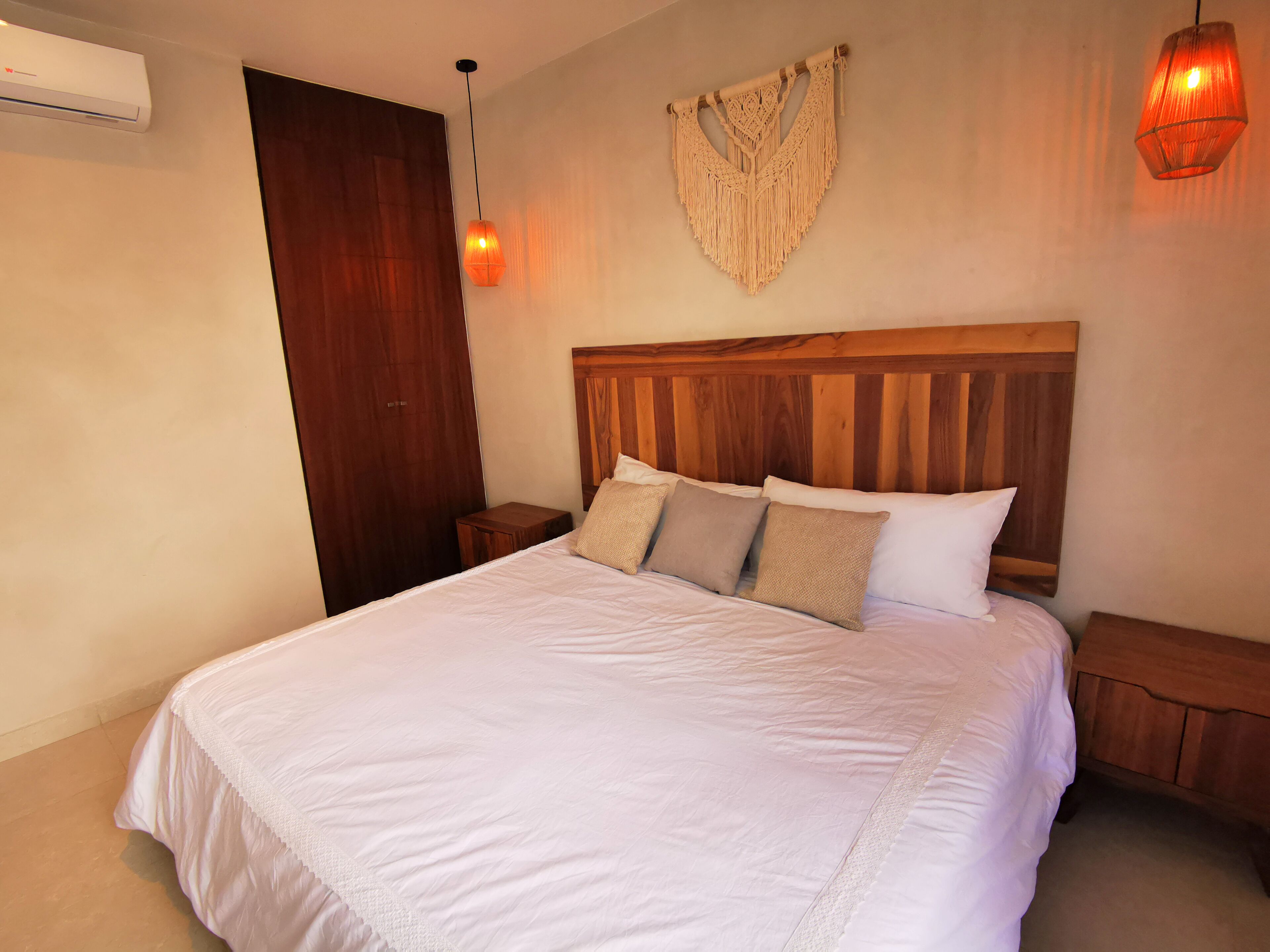 Ein ordentlich bezogenes Bett mit Holz-Kopfstück, einer an der Wand befestigten Makramee-Leuchte und einem Nachttisch mit Lampe.