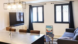 A modern living room with a sofa, a dining table with chairs, a TV, and large windows with curtains. - Duplex Standing de 77m2 Avec Rooftop de vue mer Situé Plein Centre de Bandol (Bandol)